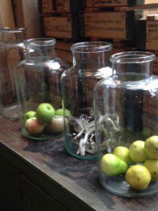 Trois bocaux en verre vintage contenant des pommes, des courges et des champignons reposent sur une étagère en bois avec des caisses empilées en arrière-plan, évoquant une charmante esthétique Brocante.