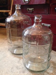 Deux grandes bonbonnes de verre vides reposent au sol, sur fond d'armoire en bois rouge, dégageant un charme rétro.