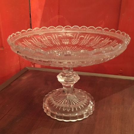La Coupe en Verre 1950 Motifs Géométriques Vendu, un plat sur piédestal en verre aux motifs ornementaux, repose sur une table en bois posée sur des panneaux de bois rouges, ajoutant une touche de charme brocante à la scène. Brocante 93