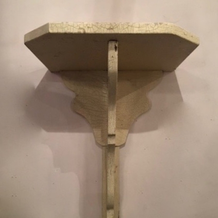 Un petit support d'étagère murale décoratif en forme de piédestal pour statue des années 1930, avec une finition de peinture craquelée de couleur crème et un charme brocante, monté contre un mur uni. Brocante 93