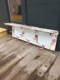 Le Porte-Manteaux & Chapeaux 1950 VENDU, avec sa finition blanche patinée et ses crochets roses, repose sur une terrasse en bois contre un mur vert foncé, dégageant une charmante ambiance vintage. Brocante 93