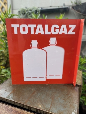 Plaque TotalGaz 1970 comportant un panneau rouge avec "TOTALGAZ" aux côtés de deux silhouettes de bonbonnes de gaz blanches, disposées en extérieur sur fond de feuillage, dégageant un charme vintage. Brocante 93