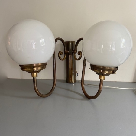 L'Applique d'Hôtel 1940 VENDU, une applique murale double vintage en laiton dotée de deux abat-jour ronds en verre blanc, se dresse gracieusement sur un fond mural beige. Brocante 93