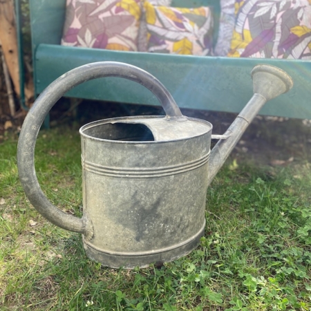 Un arrosoir en métal patiné Arrosoir Galva 1960 VENDU sur l'herbe avec un banc bleu sarcelle et des coussins colorés en arrière-plan, dégageant une charmante sensation vintage. Brocante 93