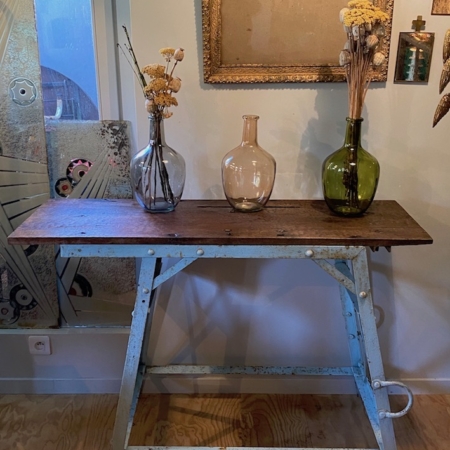 La table Etabli Industriel 1950 VENDU, une table en bois avec pieds en métal, présente trois vases remplis de fleurs séchées et est adossée à un mur décoré d'œuvres d'art encadrées, créant une ambiance vintage. Brocante 93