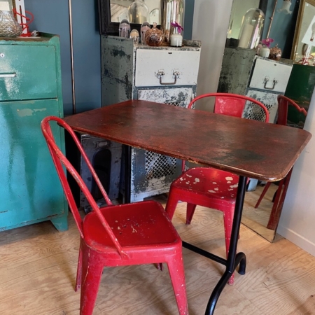 Une Table de Guinguette Pliante vintage 1950 VENDU accompagnée de deux chaises rouges vieillies trône dans une pièce ornée d'un décor rétro et d'un grand miroir, évoquant une esthétique brocante 93. Brocante 93