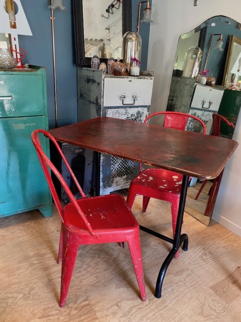Une Table de Guinguette Pliante vintage 1950 VENDU accompagnée de deux chaises rouges vieillies trône dans une pièce ornée d'un décor rétro et d'un grand miroir, évoquant une esthétique brocante 93. Brocante 93