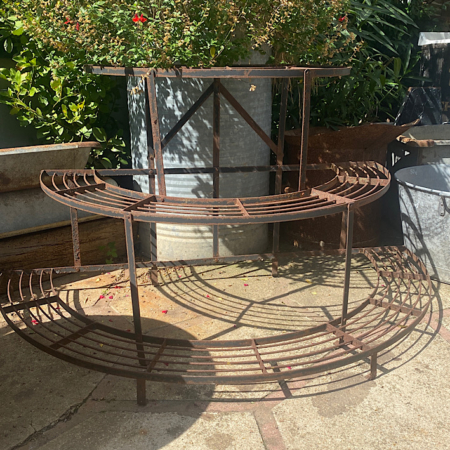 Un « Présentoir à Plantes 1960 VENDU » circulaire et rouillé est exposé à l'extérieur sur un patio en pierre, entouré de plantes vertes luxuriantes, dégageant une charmante ambiance vintage. Brocante 93