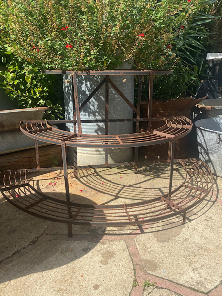 Un « Présentoir à Plantes 1960 VENDU » circulaire et rouillé est exposé à l'extérieur sur un patio en pierre, entouré de plantes vertes luxuriantes, dégageant une charmante ambiance vintage. Brocante 93