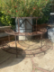 Un « Présentoir à Plantes 1960 VENDU » circulaire et rouillé est exposé à l'extérieur sur un patio en pierre, entouré de plantes vertes luxuriantes, dégageant une charmante ambiance vintage. Brocante 93