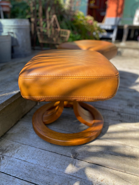 Une Paire de Poufs Cuir 1980 VENDU, avec son revêtement en cuir couleur caramel et sa base en bois, repose gracieusement sur une terrasse en bois, projetant des ombres qui rehaussent son charmante ambiance vintage. Brocante 93