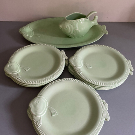 Ensemble de quatre assiettes vertes en forme de poisson avec un plat de service et une saucière assortis en forme de poisson sur une surface grise, dégageant un charme vintage parfait pour ajouter une touche d'élégance rétro à votre table, l'ensemble Service à Poisson 1950 Vendu est idéal pour toutes les occasions. Brocante 93