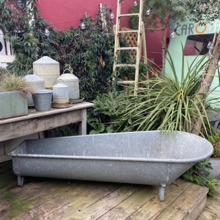 Une Baignoire Galva 1960 scintille sur une terrasse en bois au milieu de diverses plantes en pot, dégageant un charme vintage. Une échelle en bois rétro s'appuie contre un mur rouge, complétant la scène pittoresque rappelant les trouvailles de brocante. Brocante 93