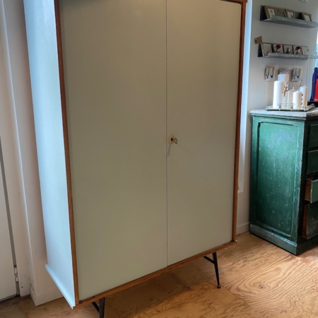 L'Armoire Penderie 1950 VENDU, un meuble vintage à deux portes et pieds élancés, trône à côté d'une commode verte dans une pièce lumineuse au plancher de bois, évoquant une charmante ambiance rétro. Brocante 93