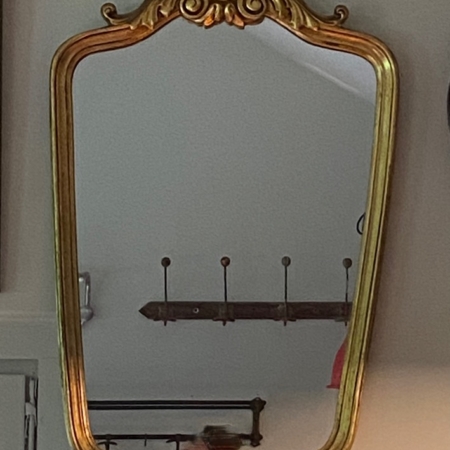 Un miroir orné au cadre doré nommé "Rococo" 1960 est accroché au mur, reflétant un porte-manteau partiellement visible et quelques objets indistincts, incarnant un charme vintage tout droit sorti de Brocante 93. Brocante 93