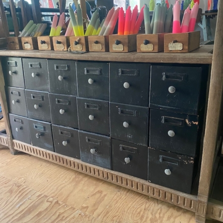 Les Tiroirs de Grainetier 1960 VENDU, un meuble en bois aux rangées de petits tiroirs, surmonté de caisses contenant des bougies colorées, dégage un charme vintage charmant. Brocante 93
