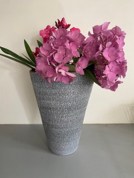 Le vase Céramique 1960, débordant de fleurs roses vibrantes et de feuilles vertes sur un fond uni, dégage un charme vintage. Brocante 93