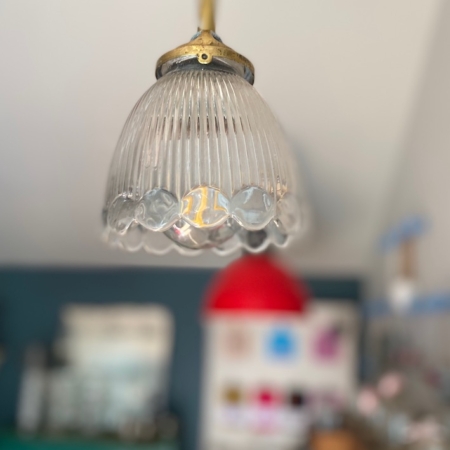 Gros plan de la Suspension Cloche Verre 1960 VENDU, une lampe suspendue vintage avec un abat-jour en verre nervuré qui évoque le charme rétro, sur fond flou comportant une lampe rouge et des étagères. Brocante 93