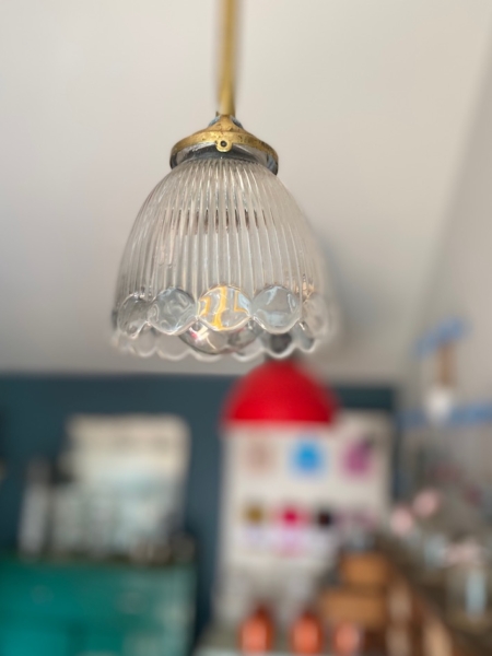 Gros plan de la Suspension Cloche Verre 1960 VENDU, une lampe suspendue vintage avec un abat-jour en verre nervuré qui évoque le charme rétro, sur fond flou comportant une lampe rouge et des étagères. Brocante 93