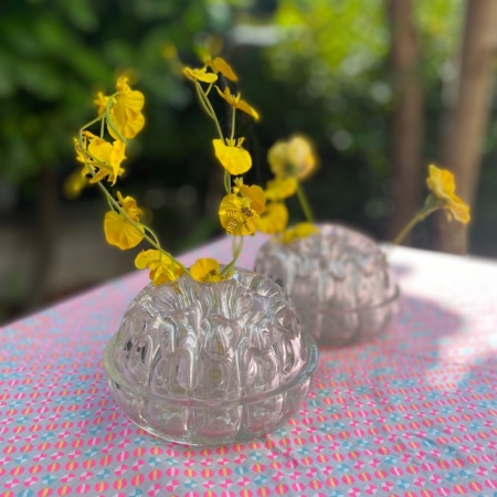 Deux vases Pique-Fleurs 1960 VENDU remplis de fleurs jaunes sur une nappe à motifs rose et blanc, sur fond de verdure, créent une charmante ambiance rétro. Brocante 93
