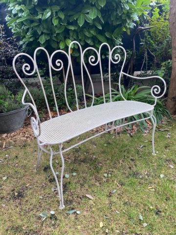 La Banquette Fer Forgé 1950 VENDU, au design de dossier orné et au charme rétro, est élégamment posée sur l'herbe dans un jardin entouré de plantes vertes et d'arbustes. Brocante 93