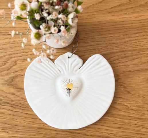 La décoration Coeur Sacré, une pièce en forme d'ange avec un coeur doré en son centre, repose sur une surface en bois patiné à côté d'un vase de petites fleurs, dégageant un charme vintage. Son esthétique brocante 93 renforce son élégance intemporelle. Brocante 93