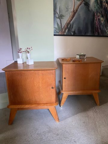 La Paire de Chevets 1960 VENDU, deux tables d'appoint rétro en bois comportant chacune une porte, sont joliment ornées de petits objets et de vases. Un tableau est suspendu au-dessus, renforçant le charme brocante 93. Brocante 93