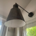 Une Applique Industrielle 1960 en métal noir, fixée au mur avec un abat-jour conique, est posée près d'une fenêtre, encadrant le feuillage vert à l'extérieur. Brocante 93