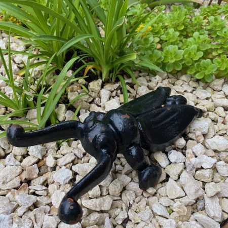 La Tire-bottes 1980 VENDU, un petit chien décoratif en céramique noire, est posée à l'envers sur un lit de gravier blanc au milieu de plantes vertes, insufflant à la scène un charme vintage. Brocante 93