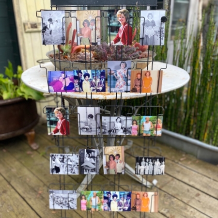 Un porte-cartes postales des années 1970, étiqueté « Porte cartes postales 1970 VENDU », est posé sur une terrasse en bois, présentant un ensemble de photographies en noir et blanc et en couleur. En arrière-plan, des plantes anciennes ajoutent au charme d'une brocante. Brocante 93
