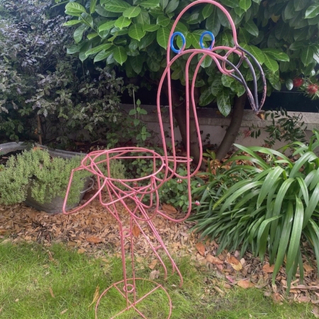 Une sculpture Flamant Rose 1960 VENDU se dresse sur l'herbe dans un jardin avec des plantes vertes et des feuilles en arrière-plan, ajoutant une touche de charme vintage. Brocante 93