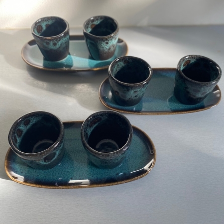 Deux ensembles de tasses à café et plateau Duo en céramique foncée sont disposés sur une surface claire, reposant sur des plateaux ovales assortis sous un éclairage doux. Brocante 93