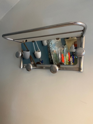 Le Porte-manteaux 1960 miroir est un porte-manteau mural en métal d'inspiration vintage, composé de cinq patères, d'une étagère et d'un miroir. Le reflet bleu du mur ajoute une touche de nostalgie, tandis que les sacs suspendus rehaussent son charme. Brocante 93