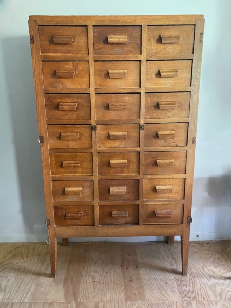 Le meuble de métier en bois 1940 est doté de 18 petits tiroirs avec poignées à bouton, posés sur un parquet contre un mur blanc, incarnant une élégance intemporelle et un charme fonctionnel. Brocante 93