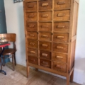 Le Meuble de métier en bois 1940, une armoire en bois vintage des années 1940, comporte plusieurs petits tiroirs et se place élégamment à côté d'une chaise et d'une table dans la pièce. Brocante 93