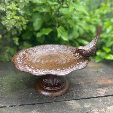 L'Abreuvoir oiseau présente une figurine d'oiseau perchée sur son rebord et est présentée sur une surface en bois avec un feuillage vert en arrière-plan. Brocante 93