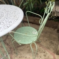 Le Salon de jardin en fer forgé 1960, doté d'une chaise et d'une table en métal vert avec des trous décoratifs, se trouve sur un patio en pierre du jardin. Brocante 93