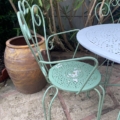 Le Salon de jardin en fer forgé 1960, doté d'une chaise en métal vert et d'une table ronde, se trouve sur un patio à côté d'un grand pot en céramique marron et de plantes à feuilles luxuriantes. Brocante 93