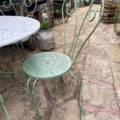 Le Salon de jardin en fer forgé 1960 présente une chaise en métal vert clair avec un dossier décoratif à côté d'une table ronde assortie, évoquant le charme vintage dans n'importe quel jardin luxuriant. Brocante 93