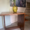 Un vase en verre vert avec des lys blancs se trouve sur la Desserte à plateaux 1940 près d'un escalier et d'une œuvre d'art de paon. Brocante 93