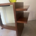 La Desserte à plateaux 1940, un meuble moderne en bois à deux étagères surmonté d'un vase vert, repose sur un sol en moquette claire. Brocante 93
