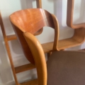 Chaise Baumann Traineau 1960 en bois avec accoudoirs courbés et assise marron, posée devant des étagères murales en bois. Brocante 93