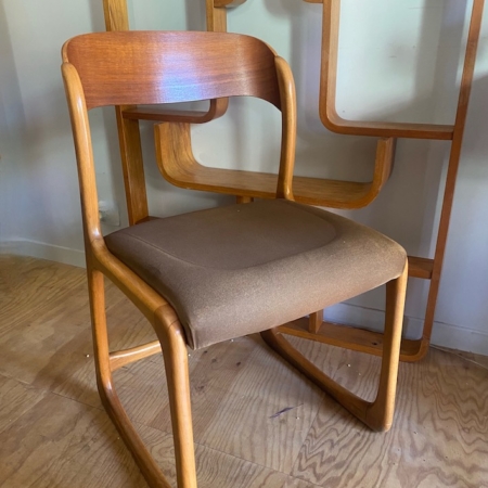 Une chaise Baumann Traineau 1960 avec une assise rembourrée marron repose sur un plancher en bois devant un panneau de bois géométrique. Brocante 93