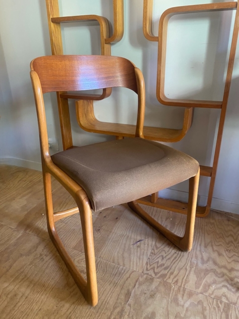Une chaise Baumann Traineau 1960 avec une assise rembourrée marron repose sur un plancher en bois devant un panneau de bois géométrique. Brocante 93