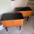 Une paire de tables de nuit 1950, au design moderne en bois du milieu du siècle avec des plateaux et des pieds noirs, se dresse sur un sol en béton près des escaliers. Brocante 93