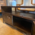 Le Meuble industriel métal riveté est un meuble TV en métal avec rangements ouverts et fermés, orné de détails rivetés. Des vases fleuris sont disposés sur le dessus, tandis que des meubles en bois se trouvent à l'arrière-plan. Brocante 93