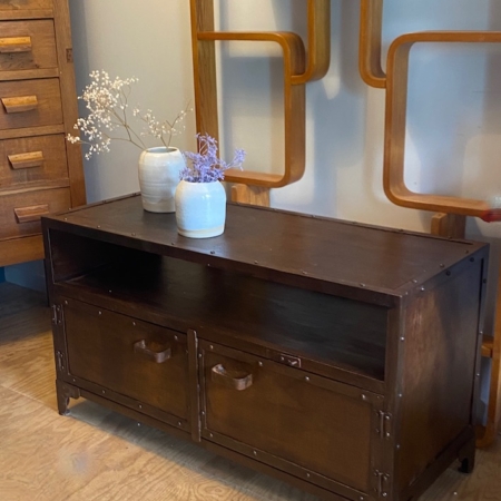 Meuble industriel métal riveté : Banc en bois foncé avec deux tiroirs, accents de rivets en métal, surmonté de deux vases en céramique remplis de fleurs et associé à un décor mural géométrique en bois. Brocante 93