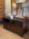 Meuble industriel métal riveté : Banc en bois foncé avec deux tiroirs, accents de rivets en métal, surmonté de deux vases en céramique remplis de fleurs et associé à un décor mural géométrique en bois. Brocante 93
