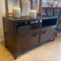Meuble industriel métal riveté : un meuble TV en bois foncé avec deux tiroirs, accents en métal riveté, deux vases de fleurs séchées et des étagères en fond—pour une touche industrielle parfaite au salon. Brocante 93