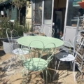 Le Salon de jardin en fer forgé 1950 présente une table et des chaises en métal vert pâle et blanc sur un patio ensoleillé, rappelant le style vintage, situé à côté de plantes luxuriantes et d'un charmant abri de jardin. Brocante 93